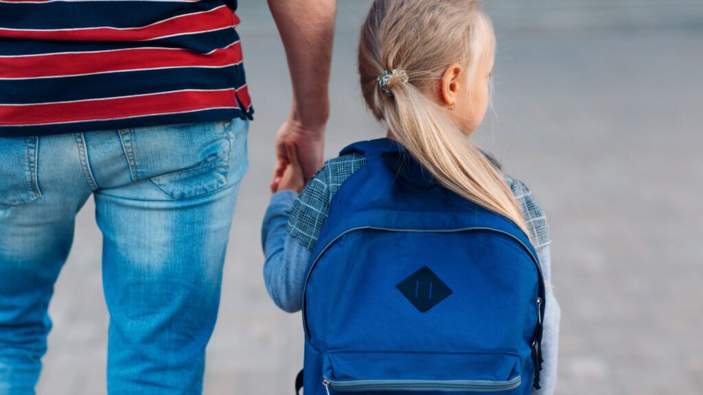 Blonde schoolgirl wearing a blue backpack and holding man's hand